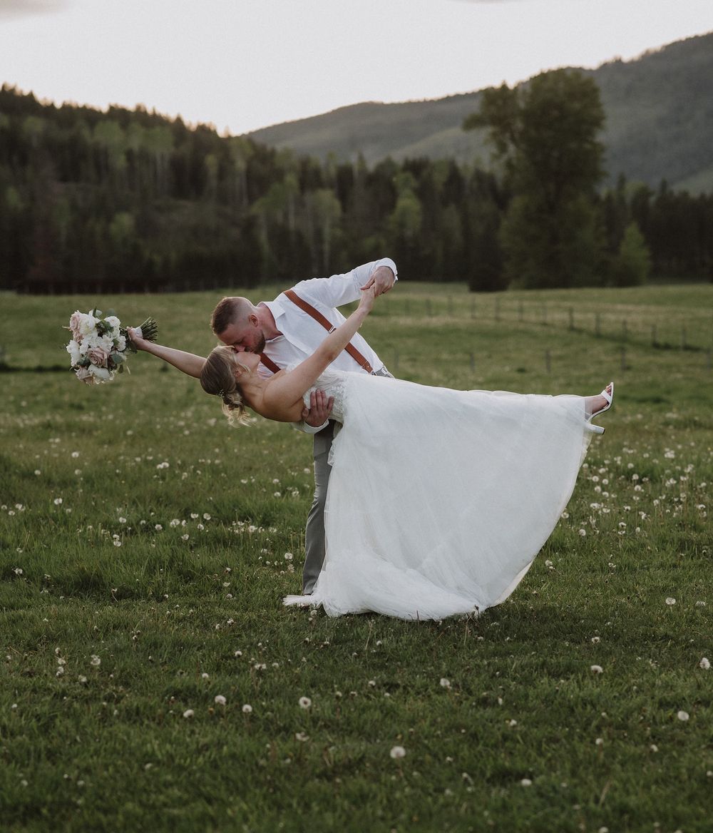 Groom dips bride in white gown for romantic kiss in lush green mountain meadow during outdoor wedding celebration.