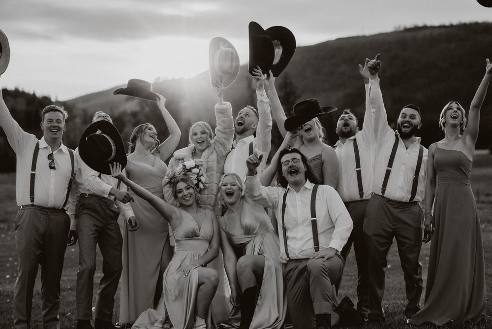 Black and white wedding party photo outdoors, group cheering with hats raised, wearing suspenders and flowing dresses.