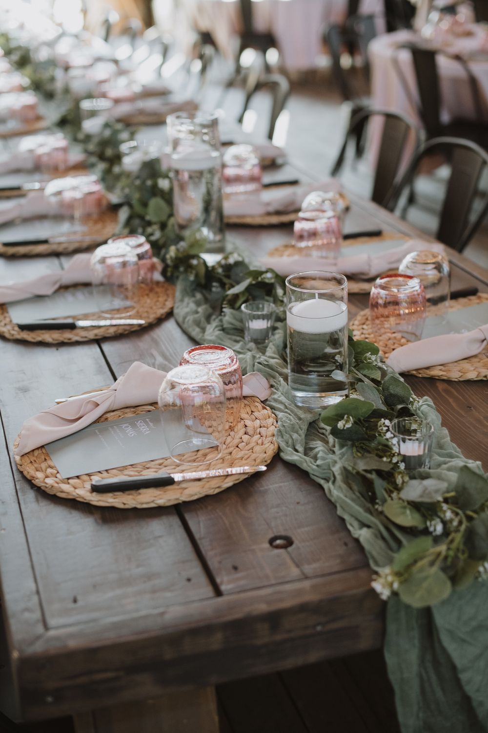Rustic wedding reception table with woven placemats, blush napkins, greenery garland runner, and floating candle centerpieces.