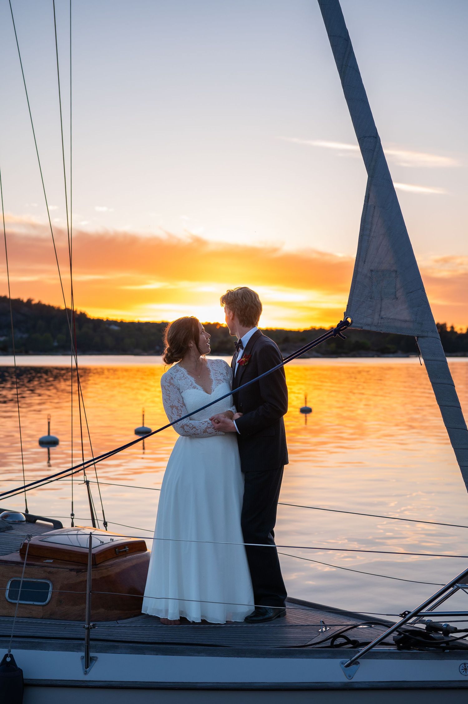 Bride and groom embrace on sailboat deck at golden sunset over calm lake on their wedding day.