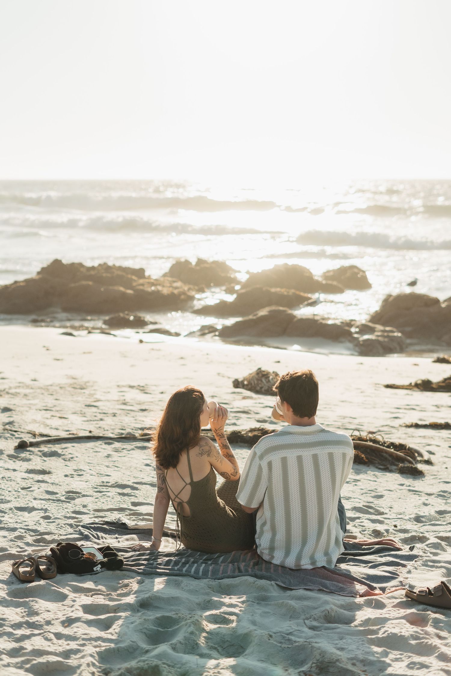Couple sitting together on sandy beach at sunset, facing ocean waves with rocky coastline in warm golden light.