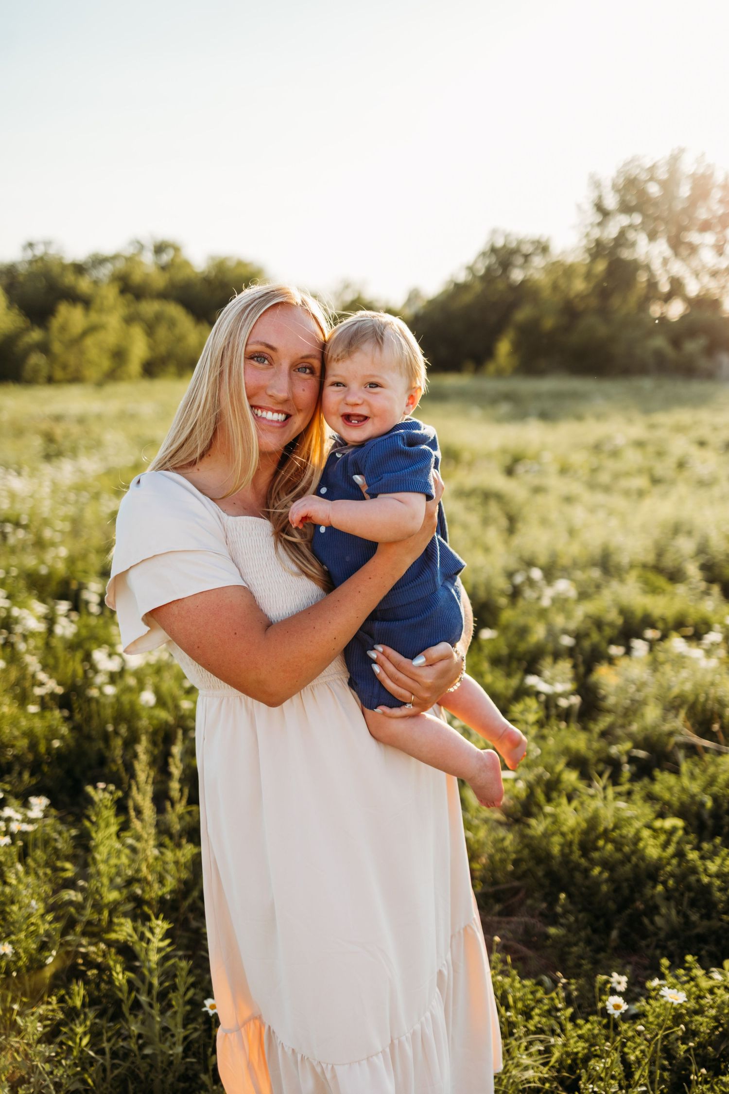 Smiling blonde woman in white dress holding a happy baby in blue outfit in a sunny green field.