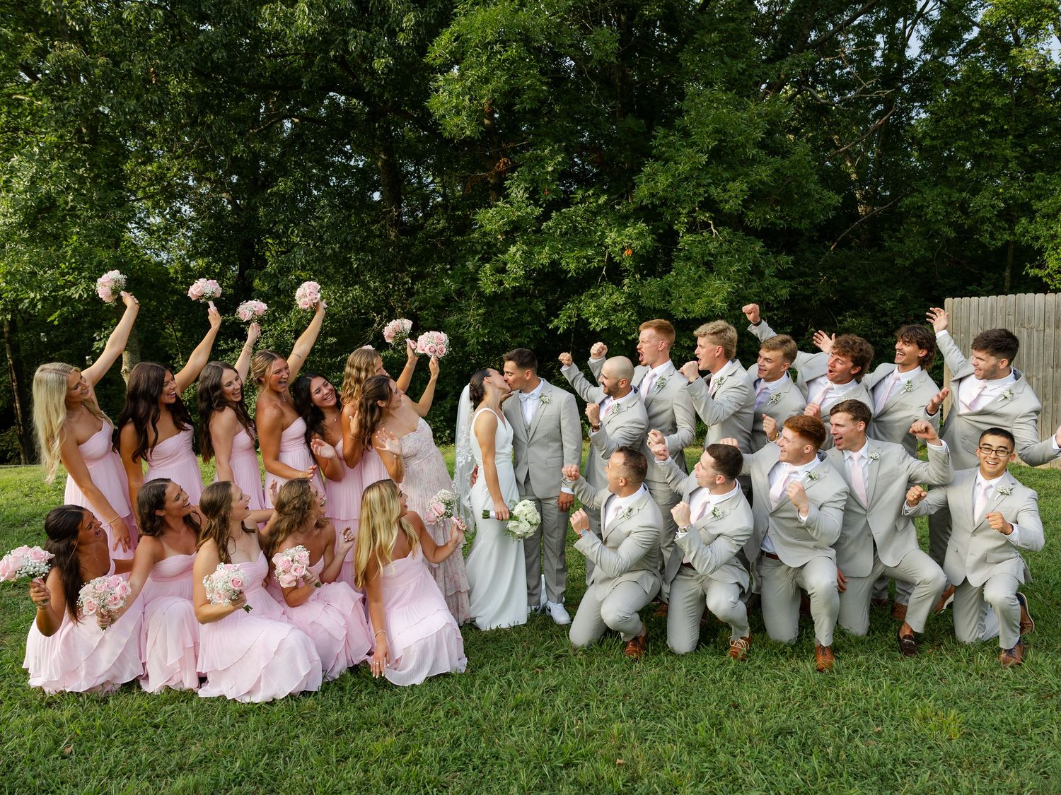 Large wedding party poses outdoors, bridesmaids in pink dresses and groomsmen in gray suits celebrating with the bride and groom.