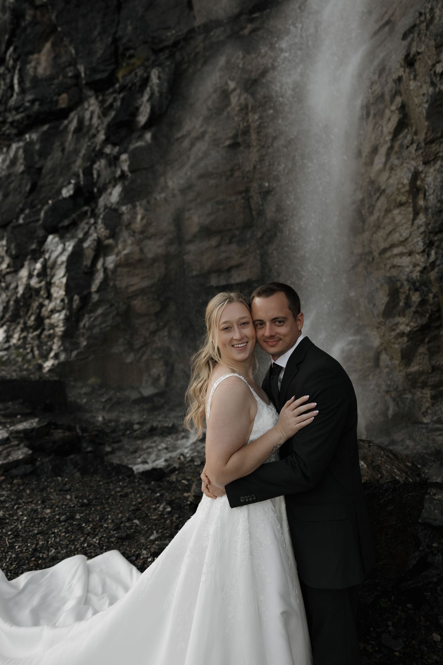 Bride in white gown with long train stands with groom in black suit on mountain overlook with rocky peaks behind them.