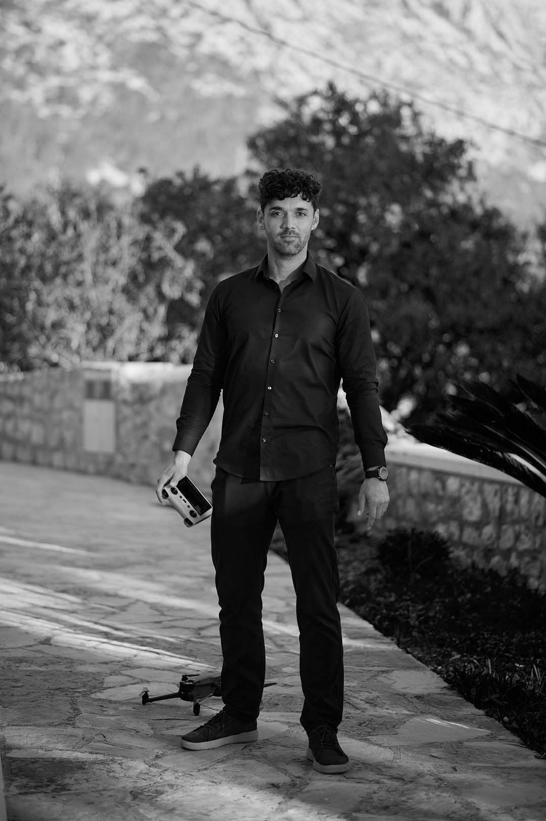 Man in navy blue shirt holding a drone controller outdoors on a stone path with green trees and mountains behind him.
