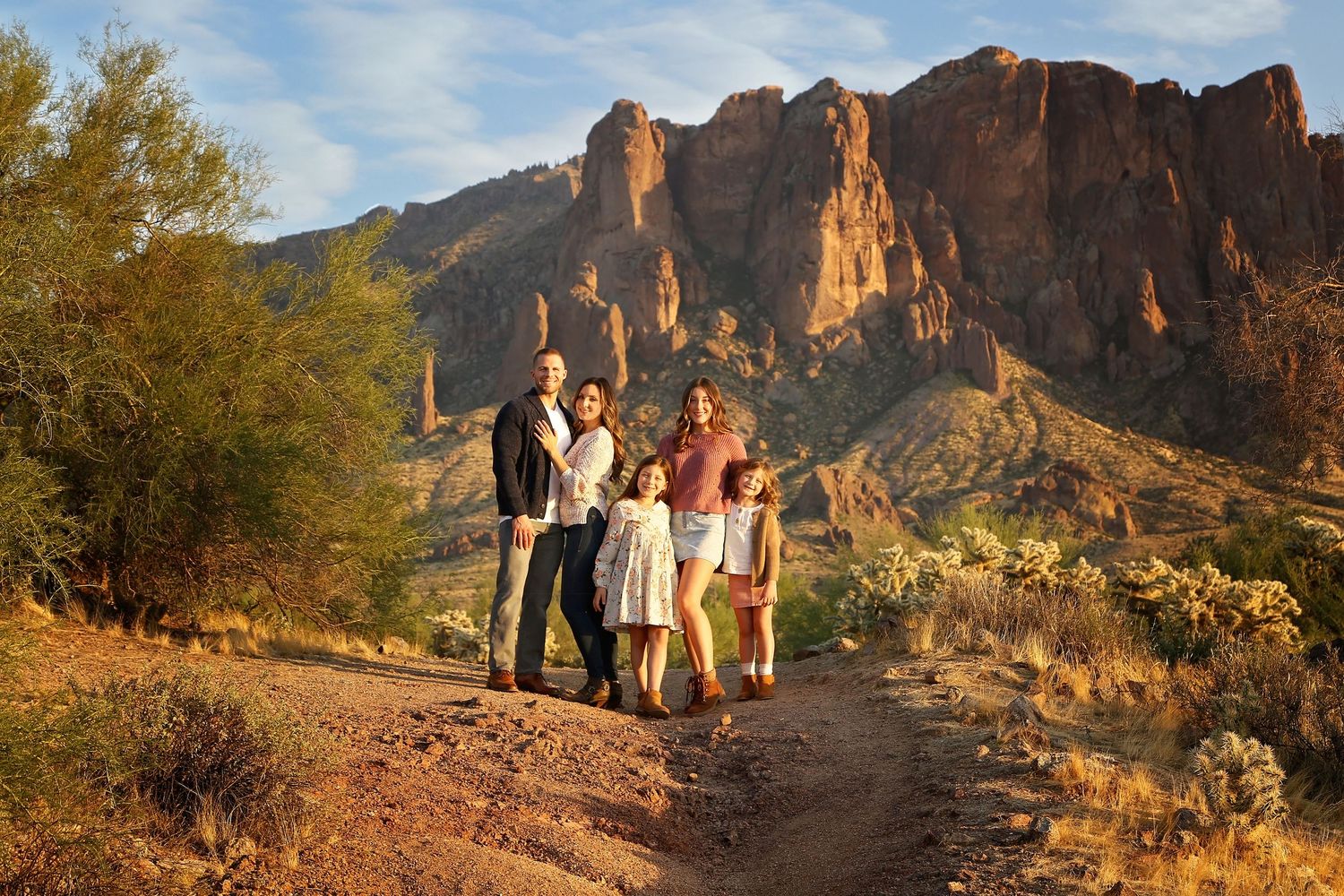 Family of five poses on a desert trail in golden light with towering red rock mountains and saguaro cacti behind them.