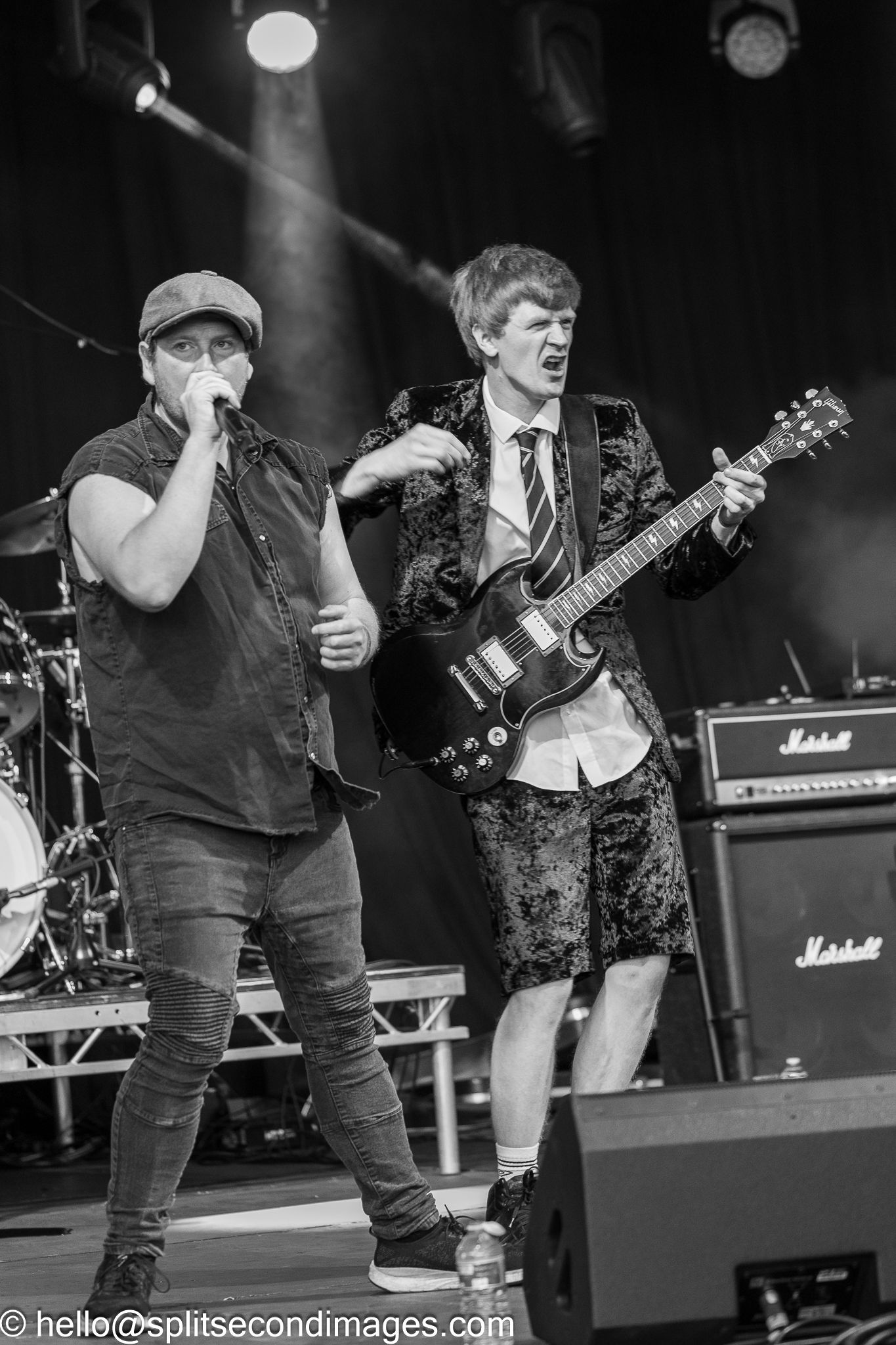 Two male performers on stage in black and white, one singing and one playing an SG guitar near Marshall amps.