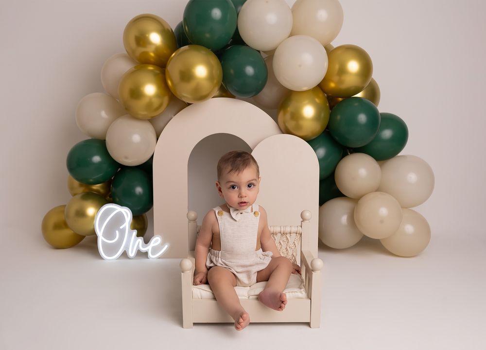 Baby sits on a mini bench surrounded by green, gold, and cream balloons with a neon “One” sign for a 1st birthday.