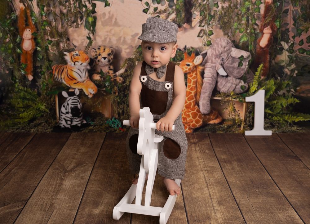 Baby in tweed overalls and cap rides a toy horse surrounded by jungle animal plush toys at a first birthday photo shoot.