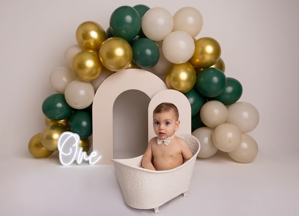 Baby sitting in a white mini bathtub with green, gold, and cream balloons and a glowing One sign for a first birthday.