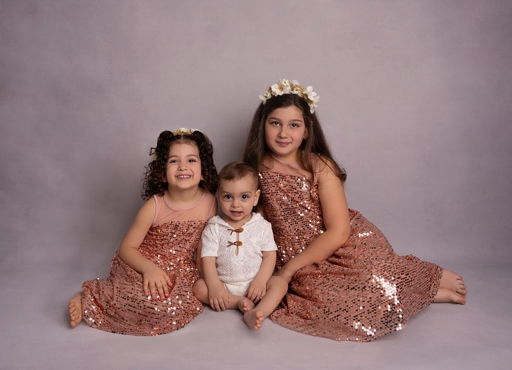 Two girls in rose gold sequin dresses pose with a baby boy in a white romper for a studio portrait.