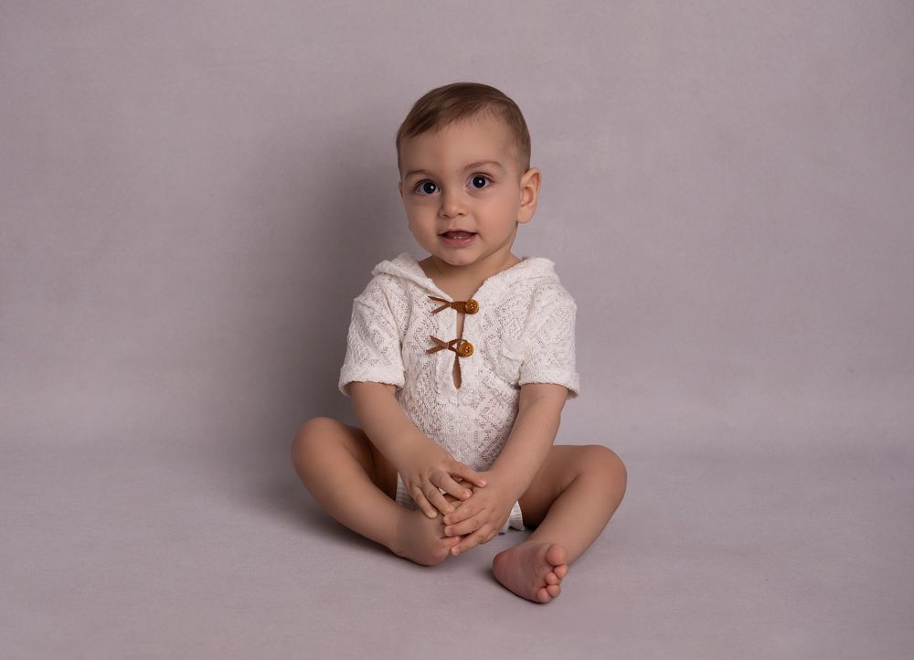Adorable baby boy in a white knit romper sits for a professional studio portrait on a gray background.