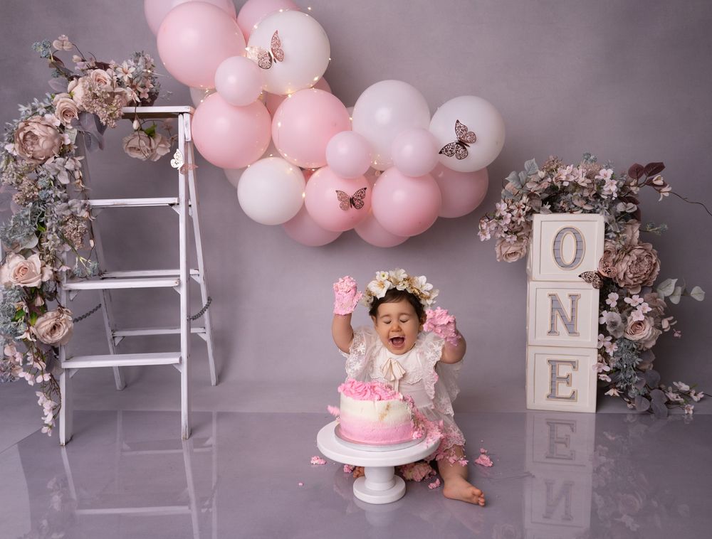 Baby girl celebrates first birthday smashing pink cake surrounded by floral arrangements, balloons, and ONE letter blocks.