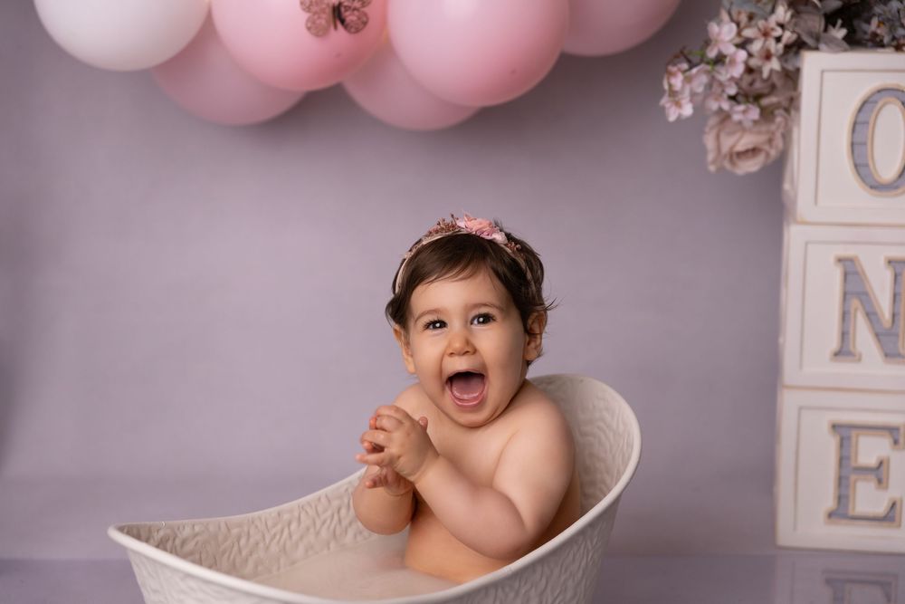 Laughing baby girl in white tub with pink balloons and ONE blocks for first birthday cake smash photo session.