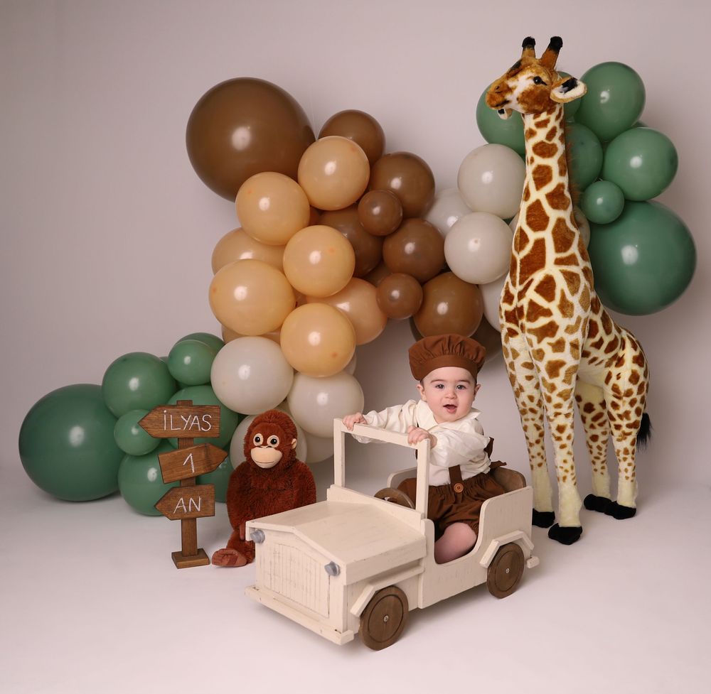 A toddler sits in a toy safari jeep surrounded by balloons, a giraffe statue, and a monkey plush at a safari birthday party.
