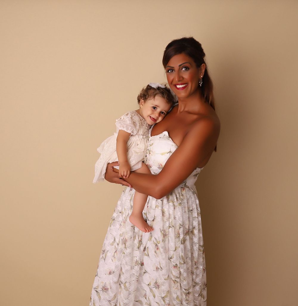 A woman in a floral dress holds a toddler girl in matching outfit against a beige studio background.