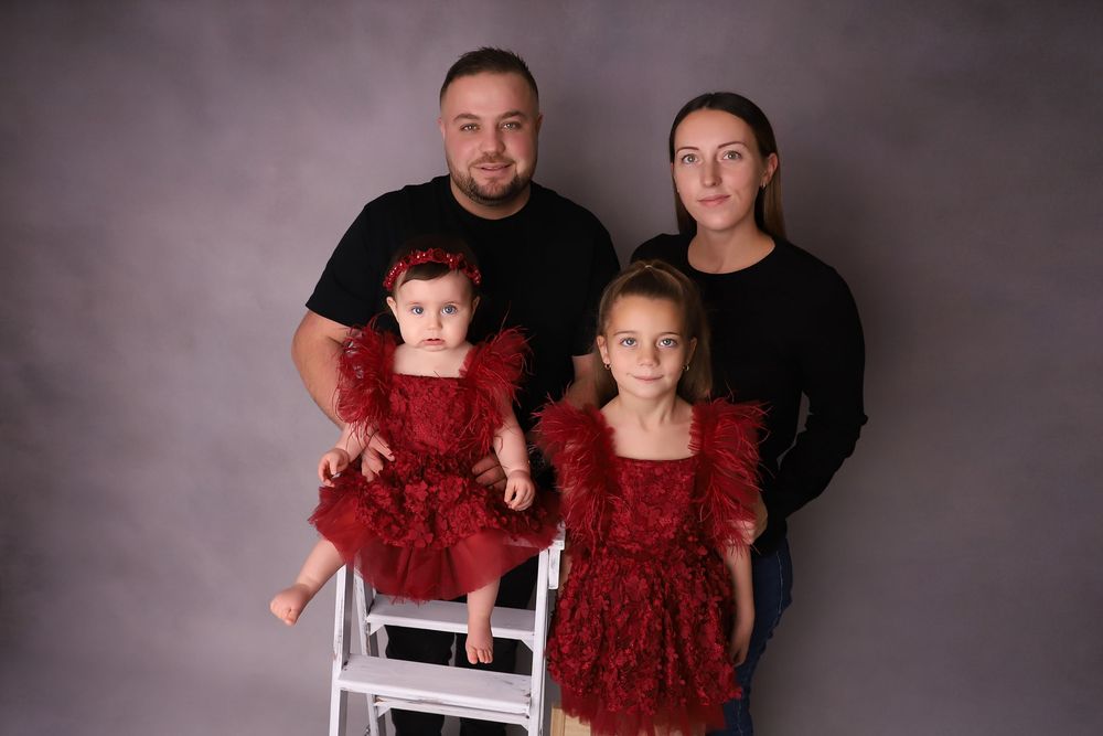 Family portrait of parents with two young girls in matching red lace dresses against a gray studio background.