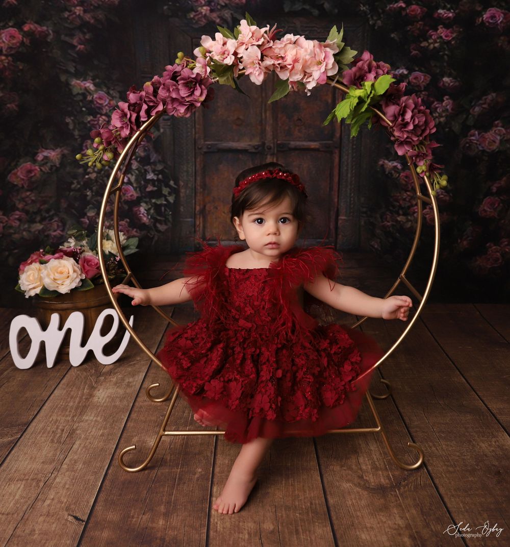 Baby girl in red lace dress sitting in gold hoop chair with flowers for her first birthday photo.