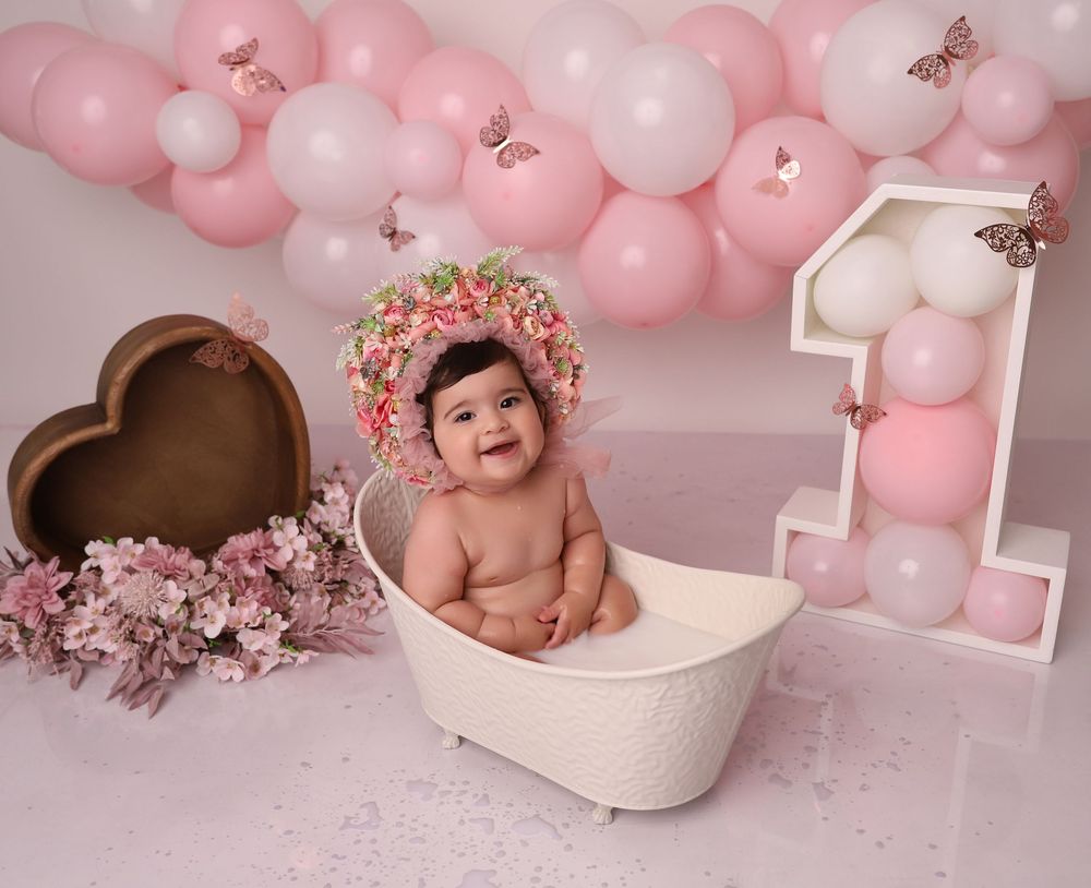 Baby girl smiling in white tub with pink balloons, butterfly decorations, floral bonnet, and number one for first birthday.