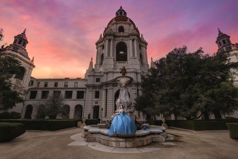A quinceañera in a blue ball gown poses by a fountain at sunset before Pasadena City Hall.