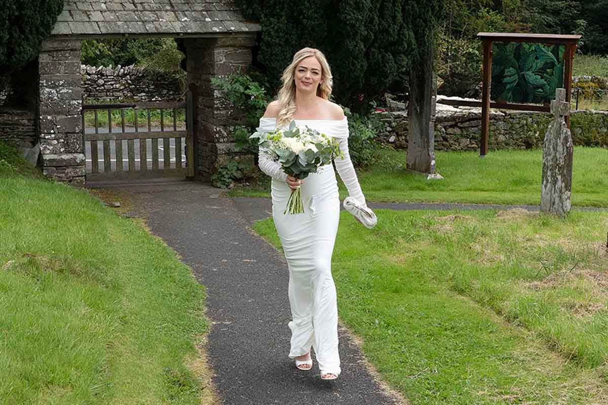 Elegant bride in white off-shoulder gown walks path carrying bouquet at her Lake District elopement wedding day