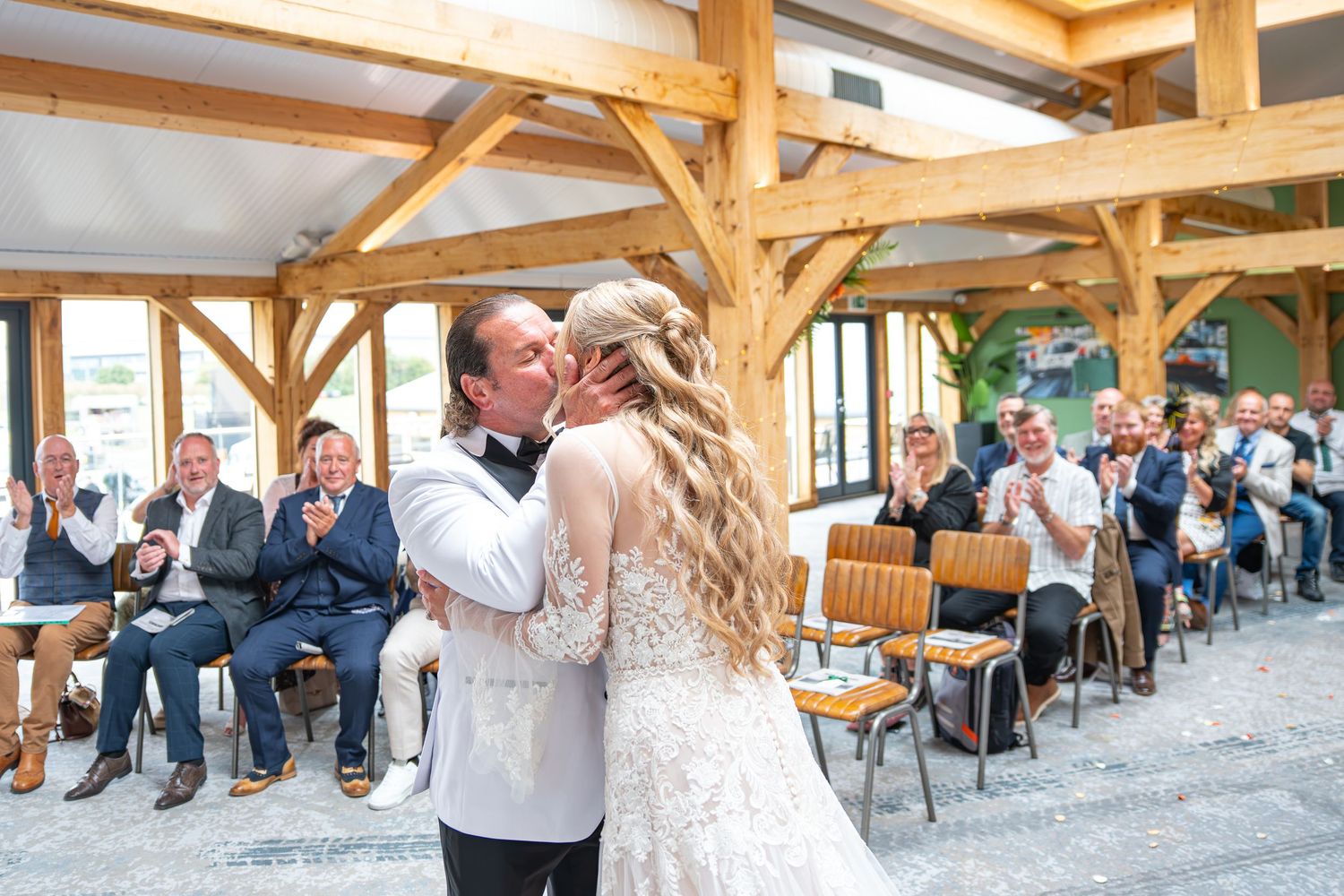 Bride and groom share their first kiss at a rustic barn wedding ceremony while guests applaud in the background.