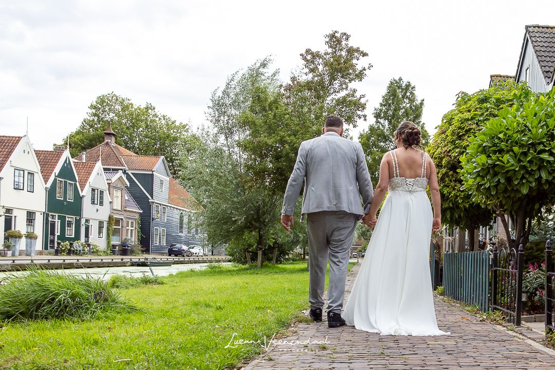 Bruid en bruidegom wandelend langs een slootje in Zaandam, we zien het bruidspar van achter. De zaanse houten huisjes staan aan de overkant.