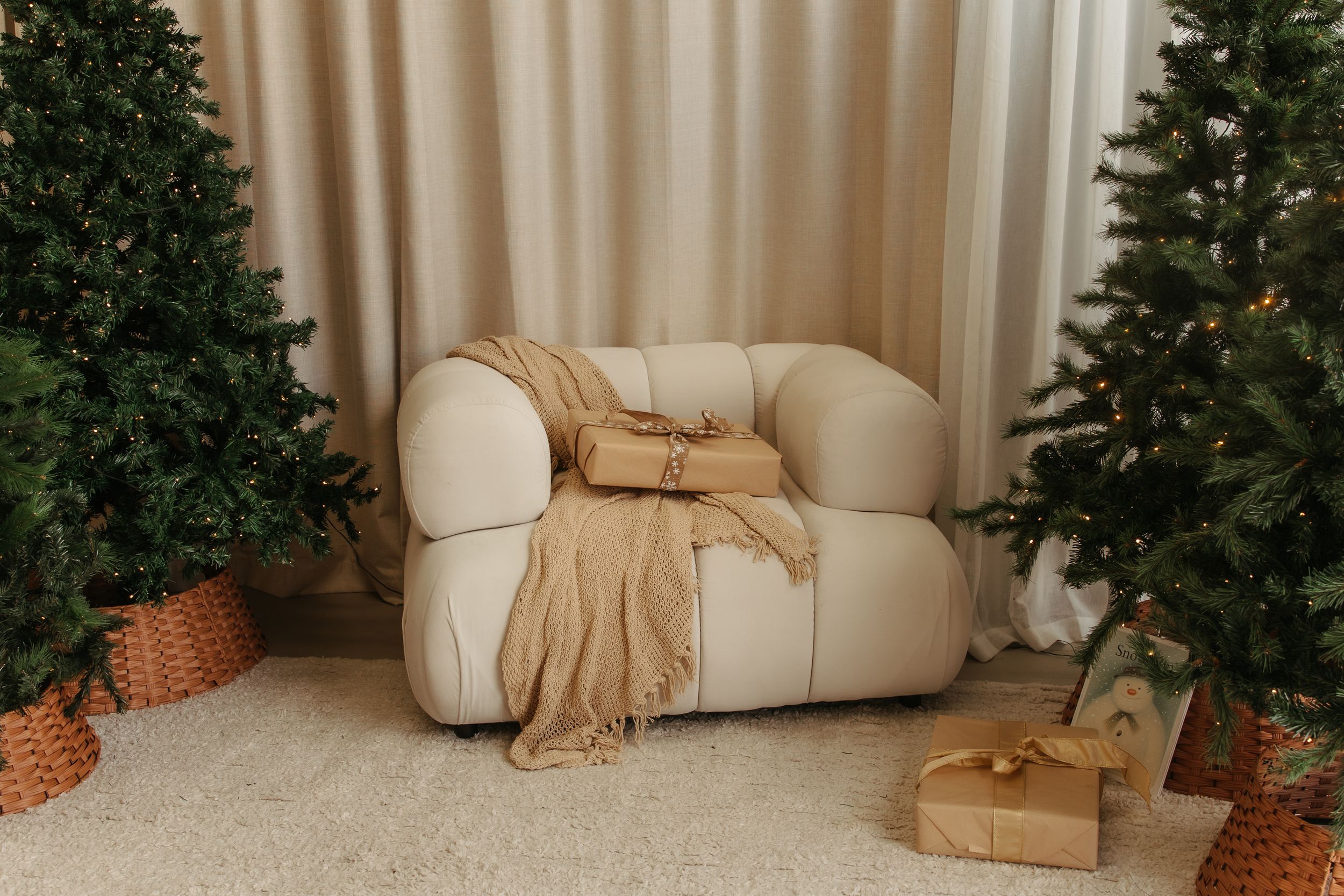 Cozy beige armchair with blanket between decorated Christmas trees and wrapped gifts on white carpet.