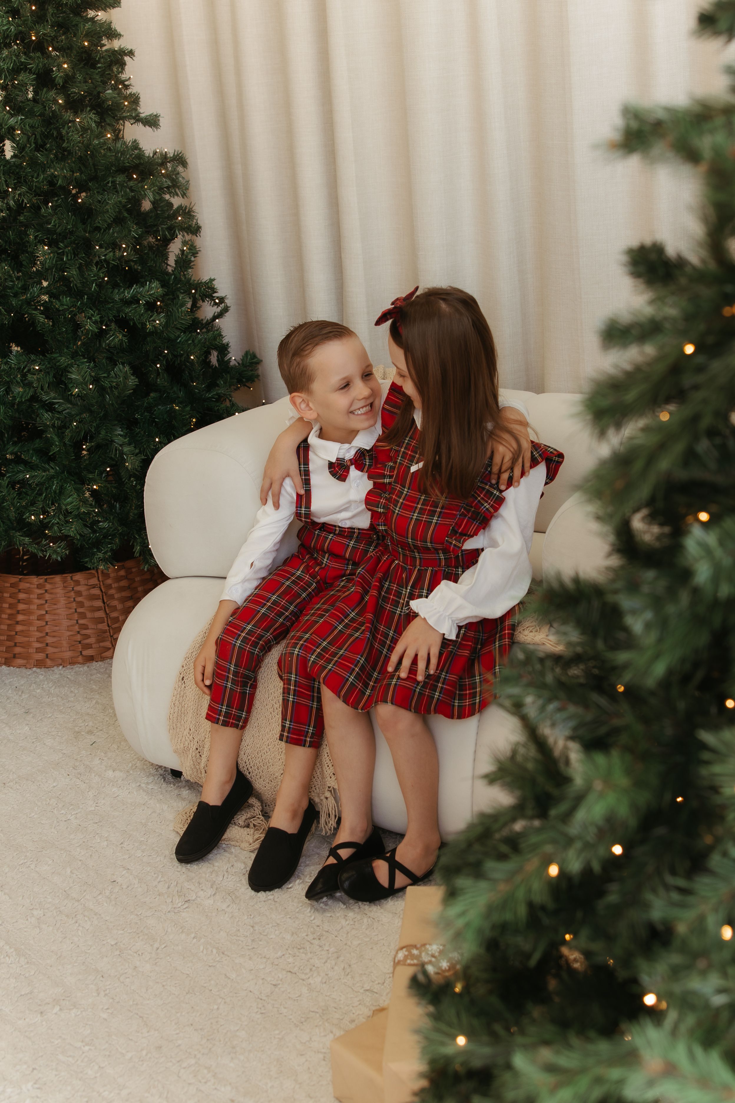 Two children in matching red plaid Christmas outfits sitting together on a white chair beside decorated Christmas trees.