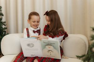 Two children in Christmas outfits reading The Snowman book together on a white sofa near a decorated tree.