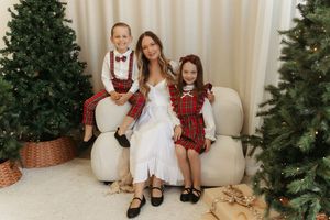 Woman in white dress with two children in red plaid outfits sitting on cream sofa between Christmas trees.