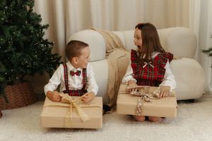 Two children in festive attire exchange Christmas gifts near a decorated tree on a white carpet.
