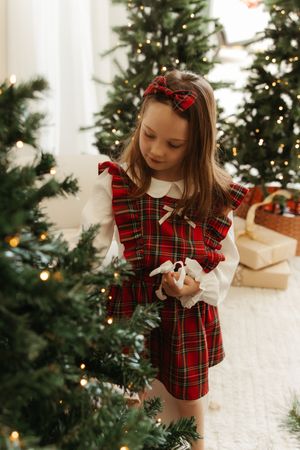 Young girl in red tartan dress decorating Christmas tree, holding toy, with festive lights and gifts in background.