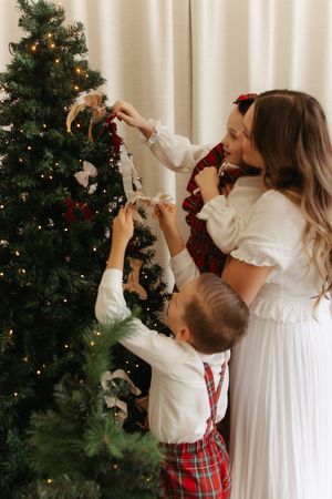 Family decorating a Christmas tree together, with mother and two children in festive plaid and white outfits.