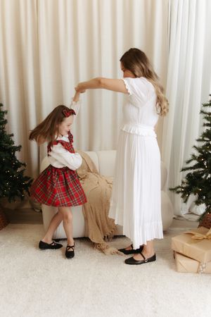 Mother in white dress twirling daughter in red plaid dress near Christmas trees with gifts.