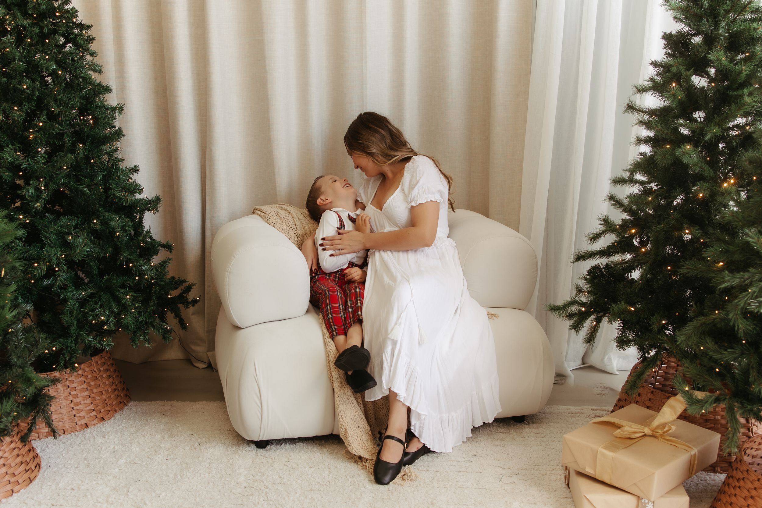 Mother and child share a tender moment on a white sofa surrounded by decorated Christmas trees and gifts.