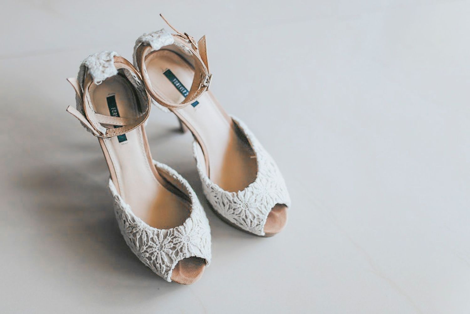Elegant white lace peep-toe wedding heels with ankle straps displayed on a light gray surface.