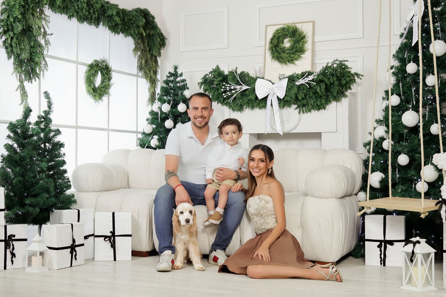 Festive living room with multiple Christmas trees, wreath, white sofa, gifts, and holiday decorations in elegant white interior.