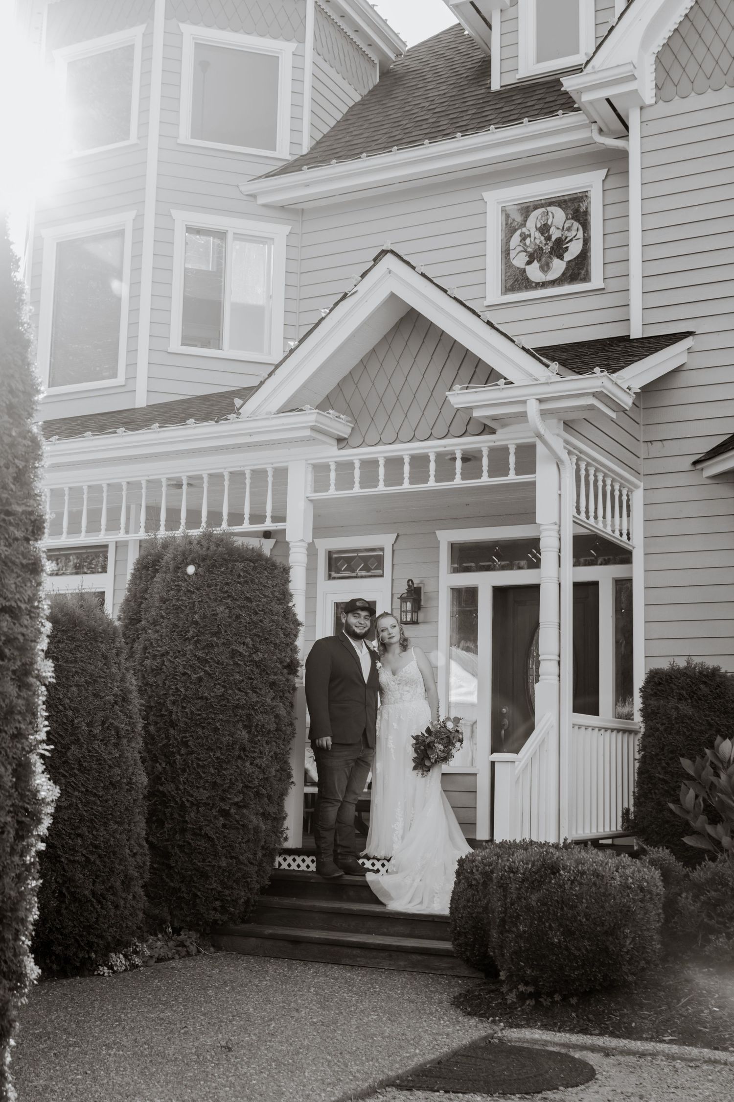 Bride in white lace gown and groom in gray jacket pose on porch steps of charming Victorian home with bouquet.