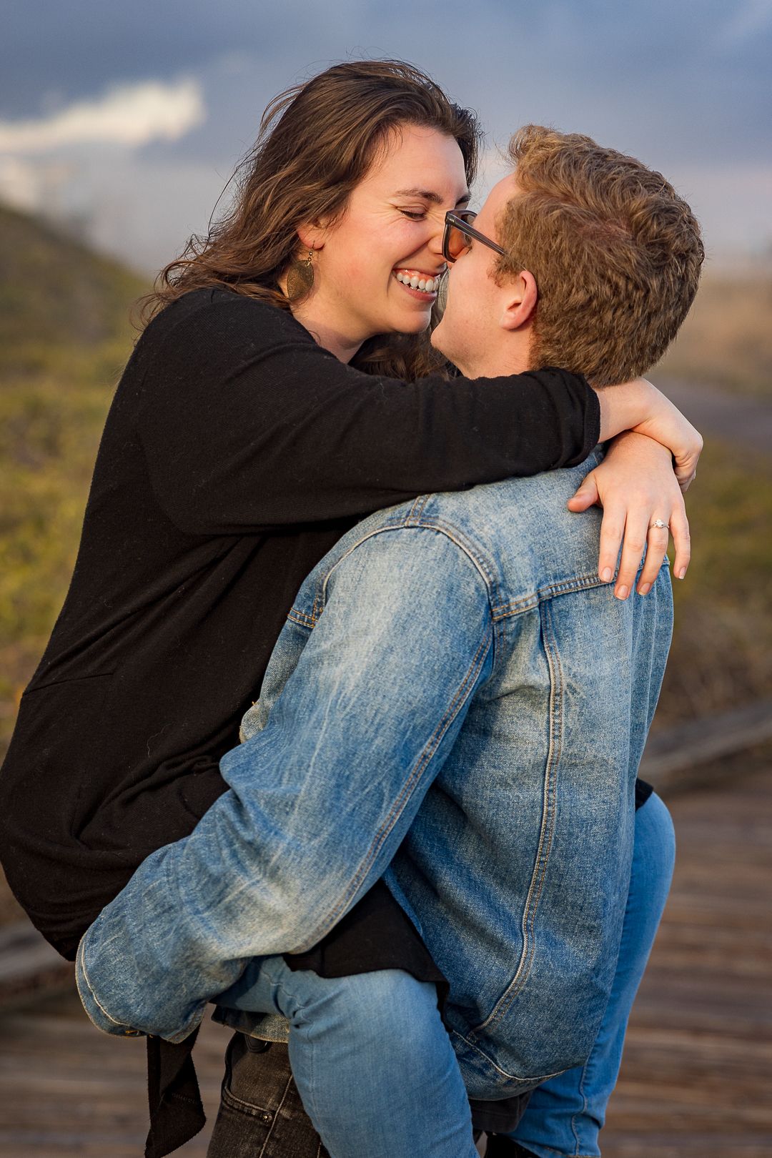 Couple sharing a joyful embrace outdoors, woman in black top laughing while being lifted by man in denim jacket.