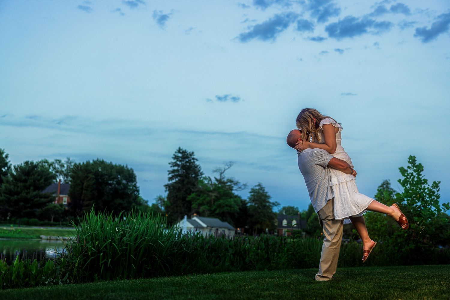 Couple sharing a romantic lift and kiss at dusk on a lush green lawn with trees and buildings in background.