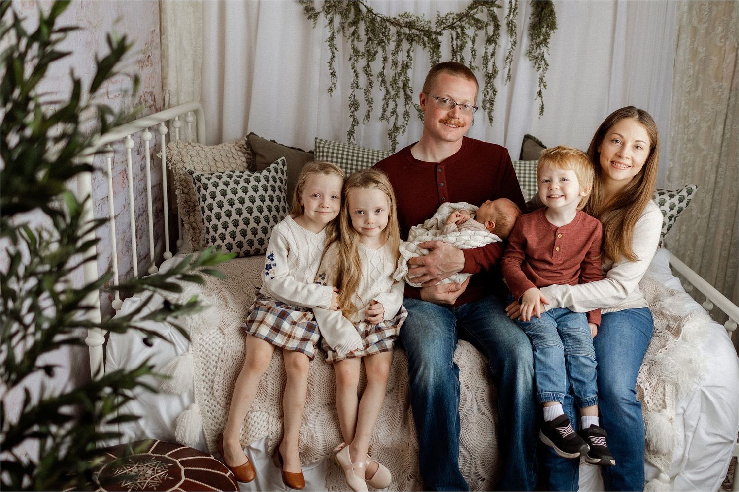 Family of six poses on a bed, parents holding newborn baby surrounded by four young children in a cozy decorated room.