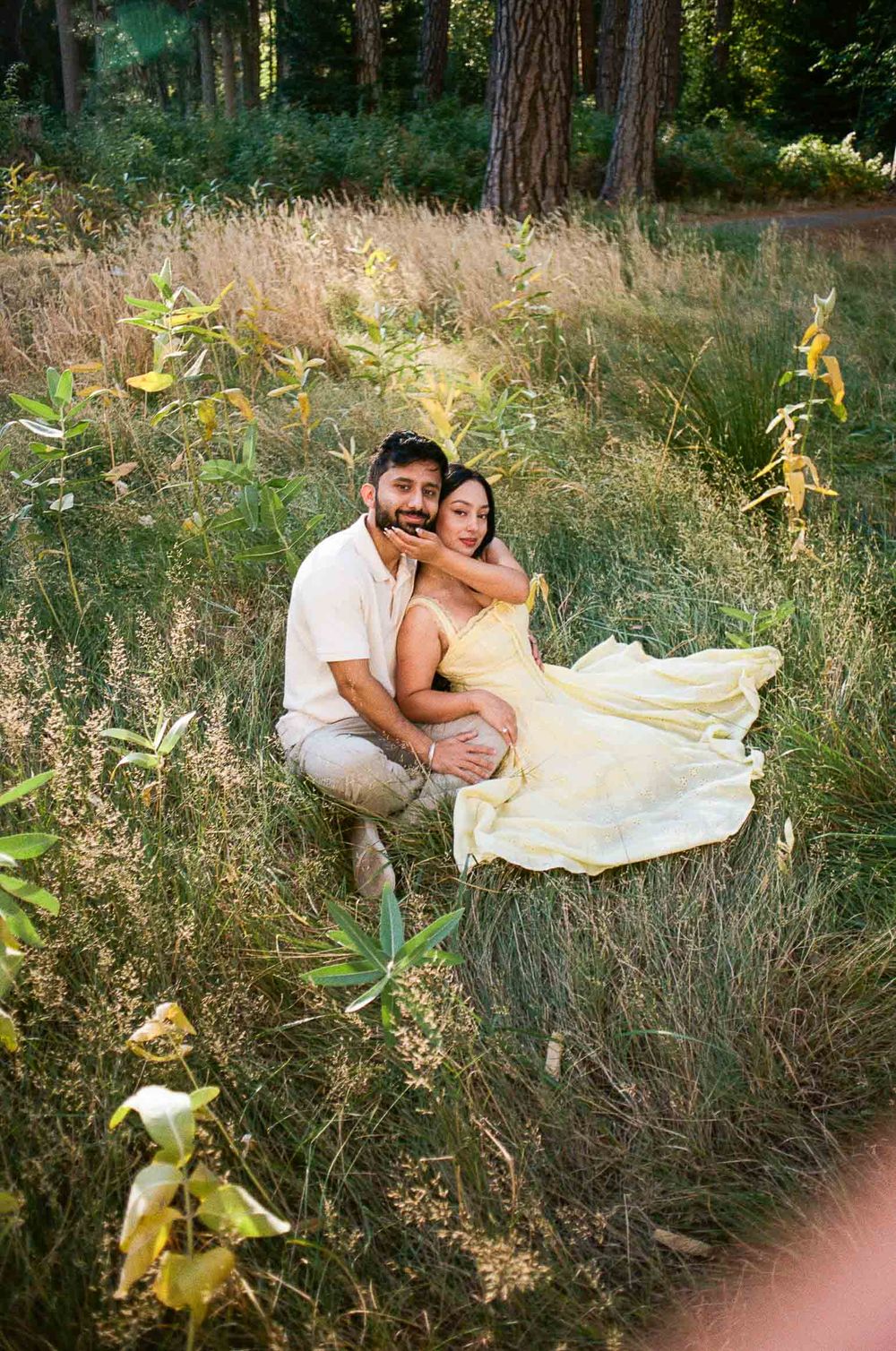 Couple sitting in a sunlit meadow surrounded by tall grass and trees, woman in yellow dress embracing man from behind.