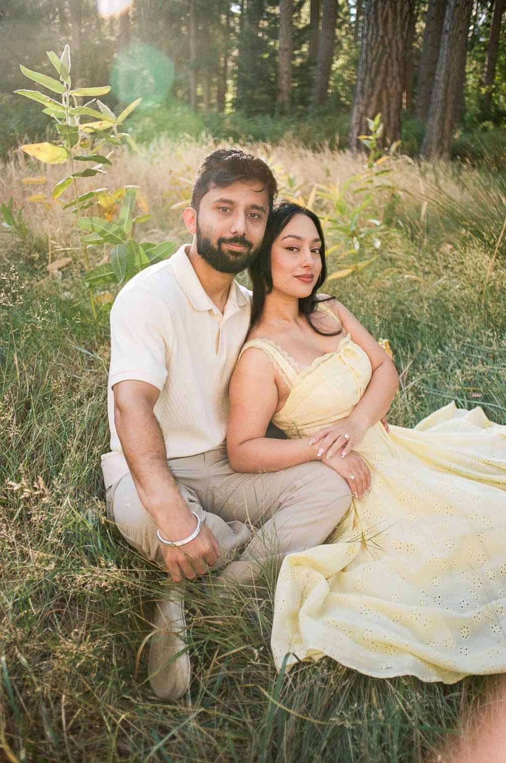 Couple sitting in golden grass field, woman in yellow dress leaning on man in cream polo shirt.