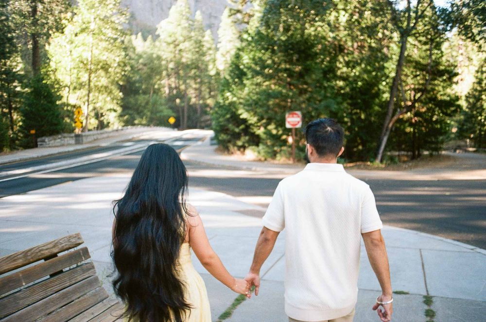 Couple holding hands walking down a road surrounded by tall green trees in Yosemite National Park.