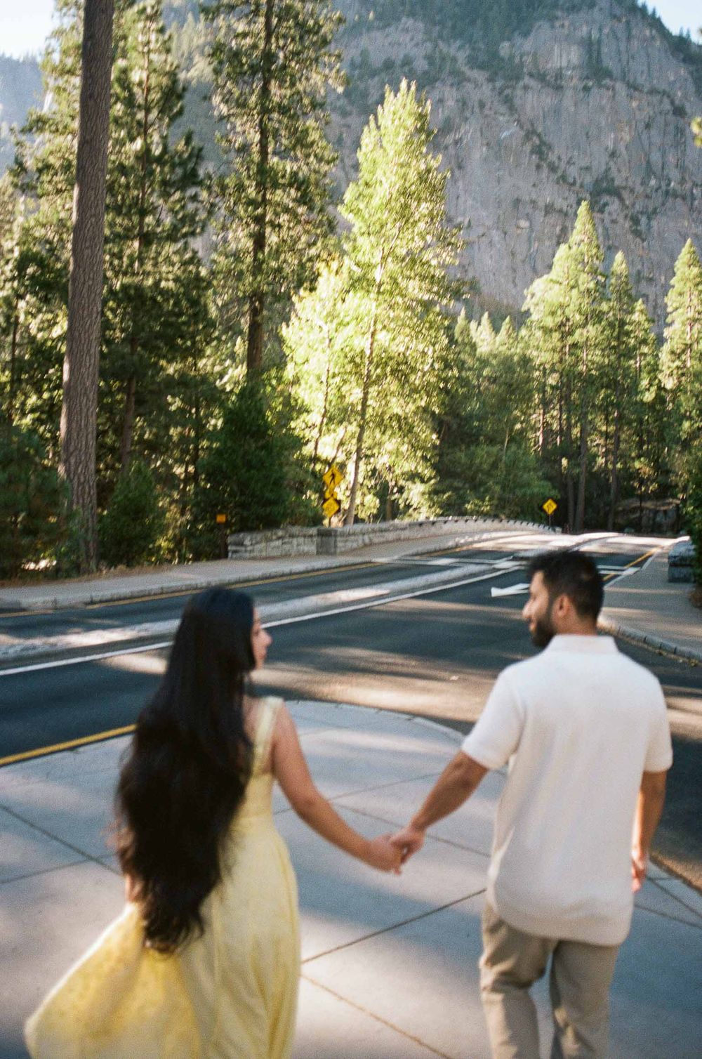 Couple holding hands walking toward a tree-lined road with a rocky mountain backdrop in Yosemite National Park.