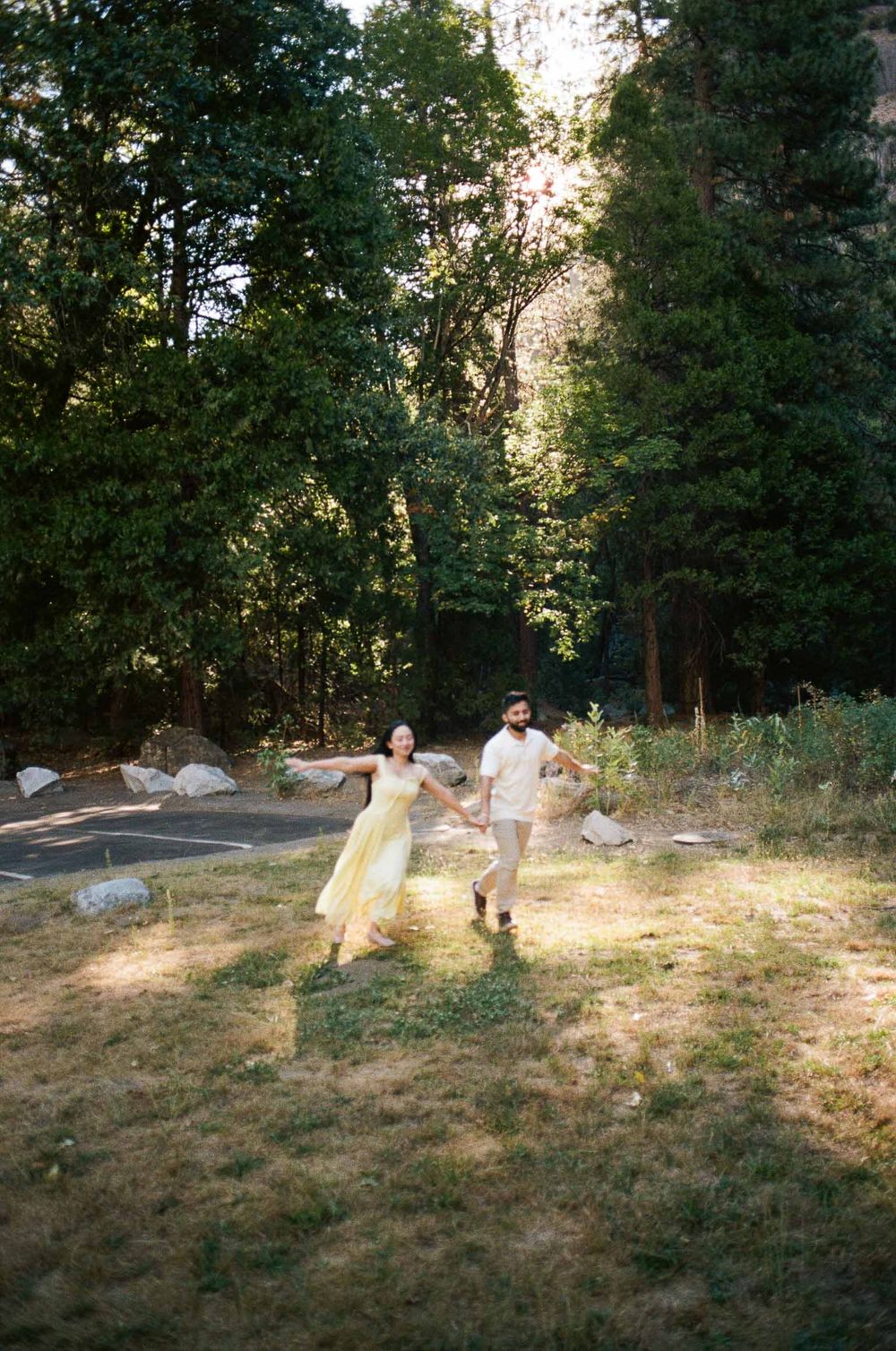 Joyful couple running hand-in-hand through sunlit Yosemite forest clearing, woman in yellow dress.