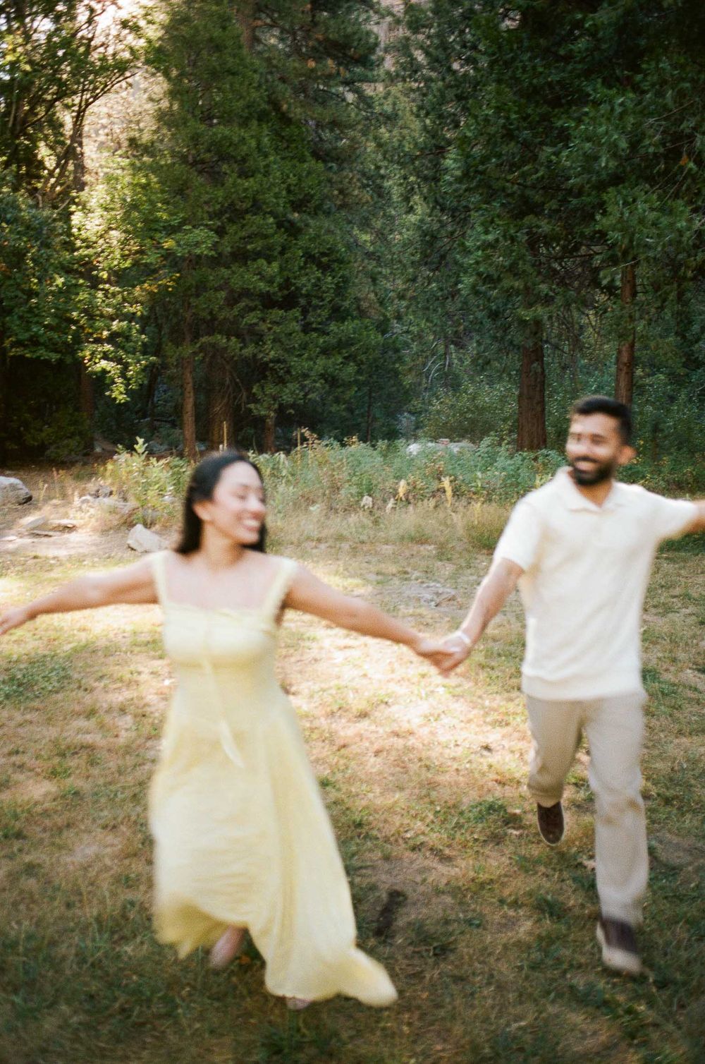 A joyful couple holds hands and runs through a sunlit forest clearing, the woman wearing a flowing yellow dress.