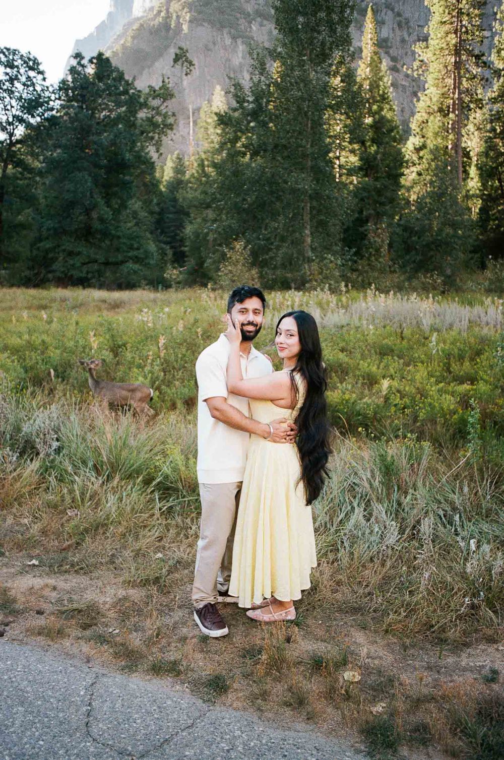 Couple embracing in Yosemite meadow with deer, tall pines, and granite cliffs in background, woman in yellow dress.