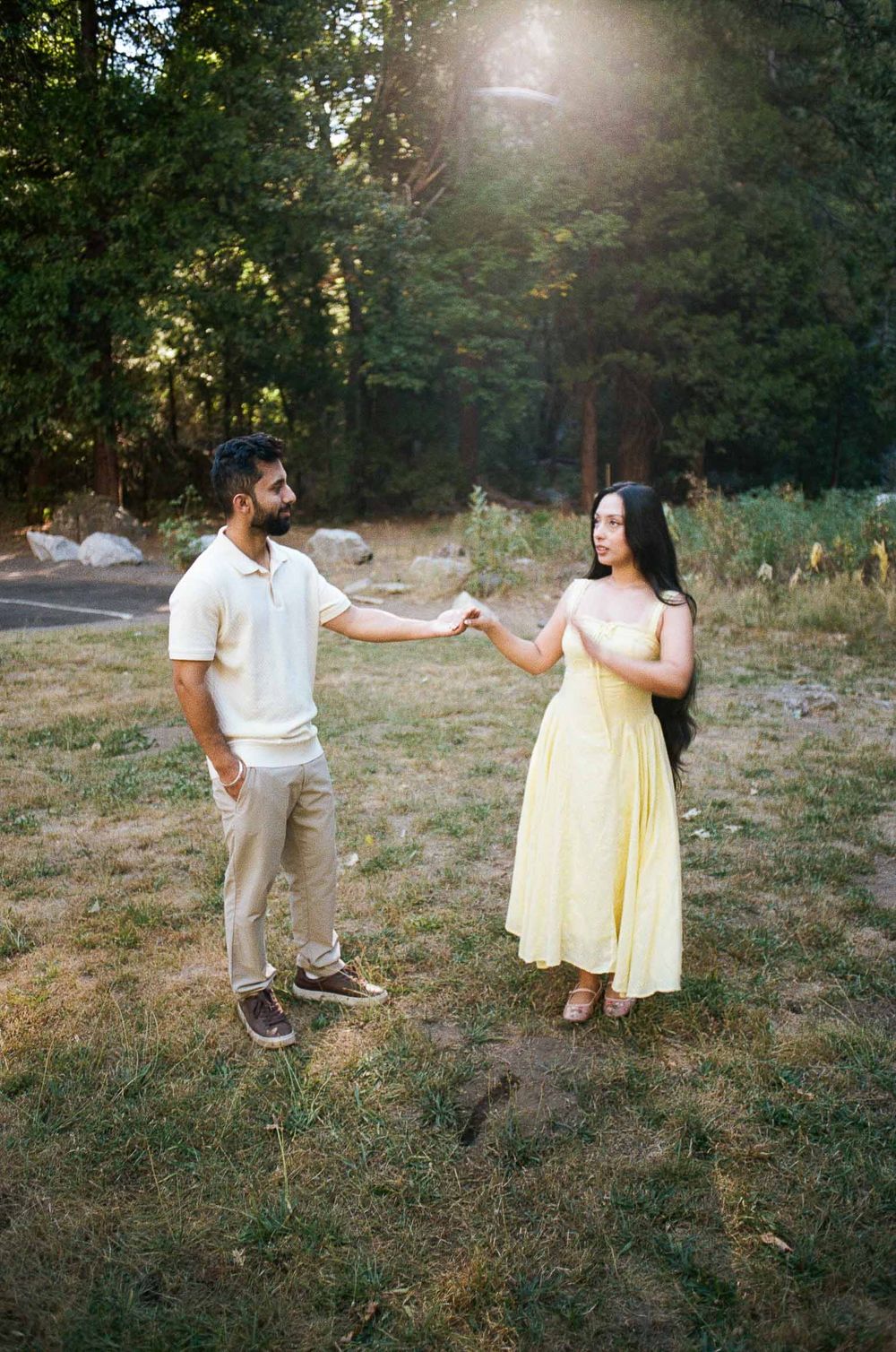 Couple holding hands in a sunlit forest clearing, man in white polo and woman in yellow maxi dress.